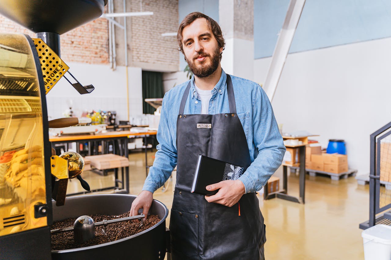 home-services-004 A bearded master roaster stands by a coffee roasting machine, holding a notebook in an industrial workshop.